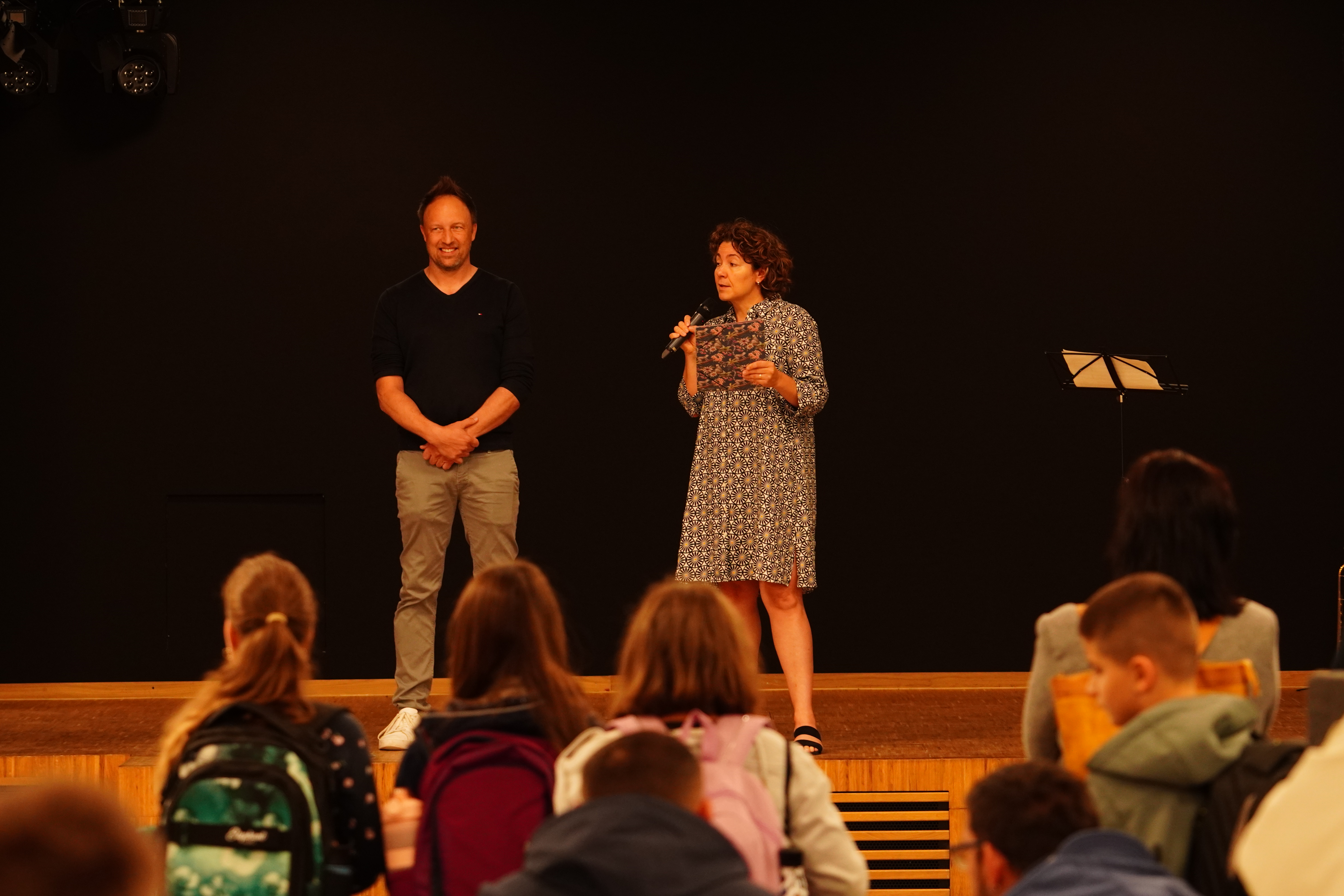 Auf dem Bild stehen Herr Patrick Hahn und Frau Claudia Gaßner auf einer Bühne. Claudia Gaßner spricht in ein Mikrofon. Vor der Bühne sieht man die Hinterköpfe der Schülerinnen und Schüler.